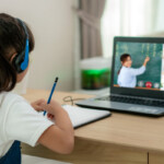 A little girl is learning online with the help of laptop and headphone besides, she has notebook to take notes and pen stand near to her.