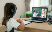 A little girl is learning online with the help of laptop and headphone besides, she has notebook to take notes and pen stand near to her.