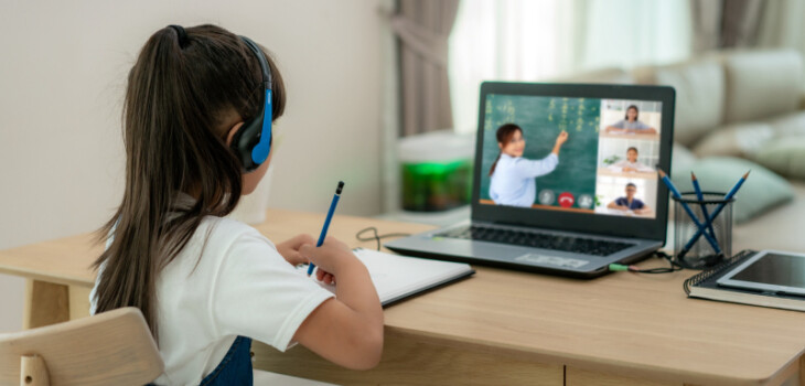 A little girl is learning online with the help of laptop and headphone besides, she has notebook to take notes and pen stand near to her.