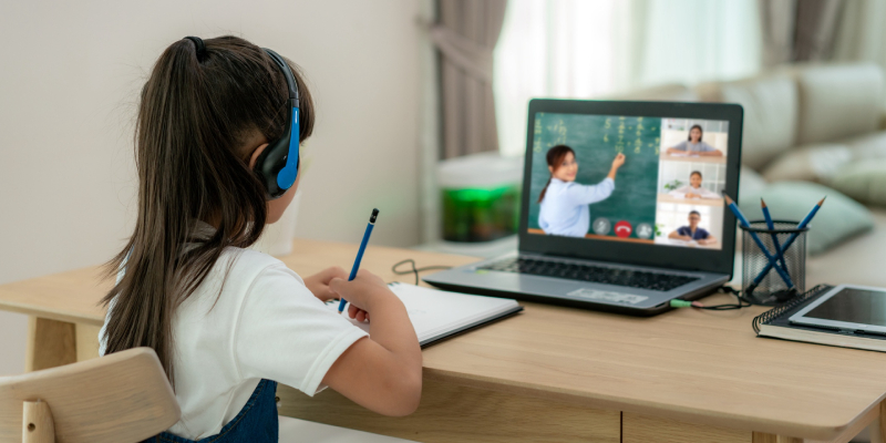 A little girl is learning online with the help of laptop and headphone besides, she has notebook to take notes and pen stand near to her.