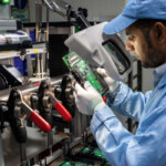 An engineer at a manufacturing industry is checking a circuit through a thick lens, we can see some other equipment around.