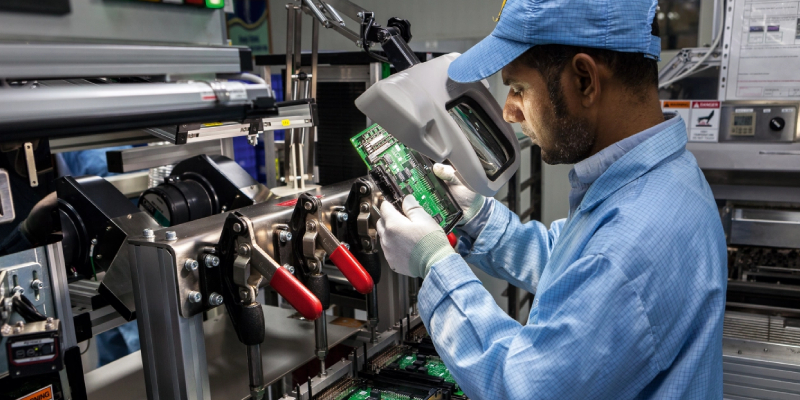 An engineer at a manufacturing industry is checking a circuit through a thick lens, we can see some other equipment around.