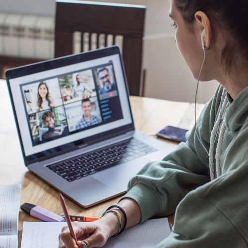 A teen-age girl is wearing a headphone, listening to her online class via laptop and taking notes on her notepad.