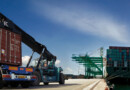 At a sea port there two heavy vehicle that is loaded with heavy cargo containers, on the other hand there is a ship in the sea that carries many containers.