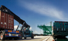 At a sea port there two heavy vehicle that is loaded with heavy cargo containers, on the other hand there is a ship in the sea that carries many containers.