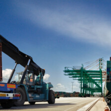At a sea port there two heavy vehicle that is loaded with heavy cargo containers, on the other hand there is a ship in the sea that carries many containers.