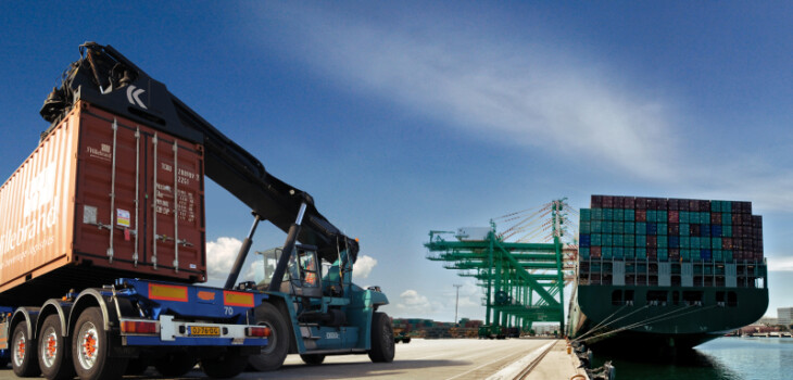 At a sea port there two heavy vehicle that is loaded with heavy cargo containers, on the other hand there is a ship in the sea that carries many containers.