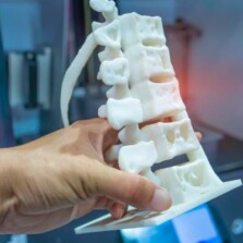 Close up mage of a persons hand holding a white 3D model of a portion of human spine inside a blurred printer room.