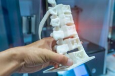 Close up mage of a persons hand holding a white 3D model of a portion of human spine inside a blurred printer room.